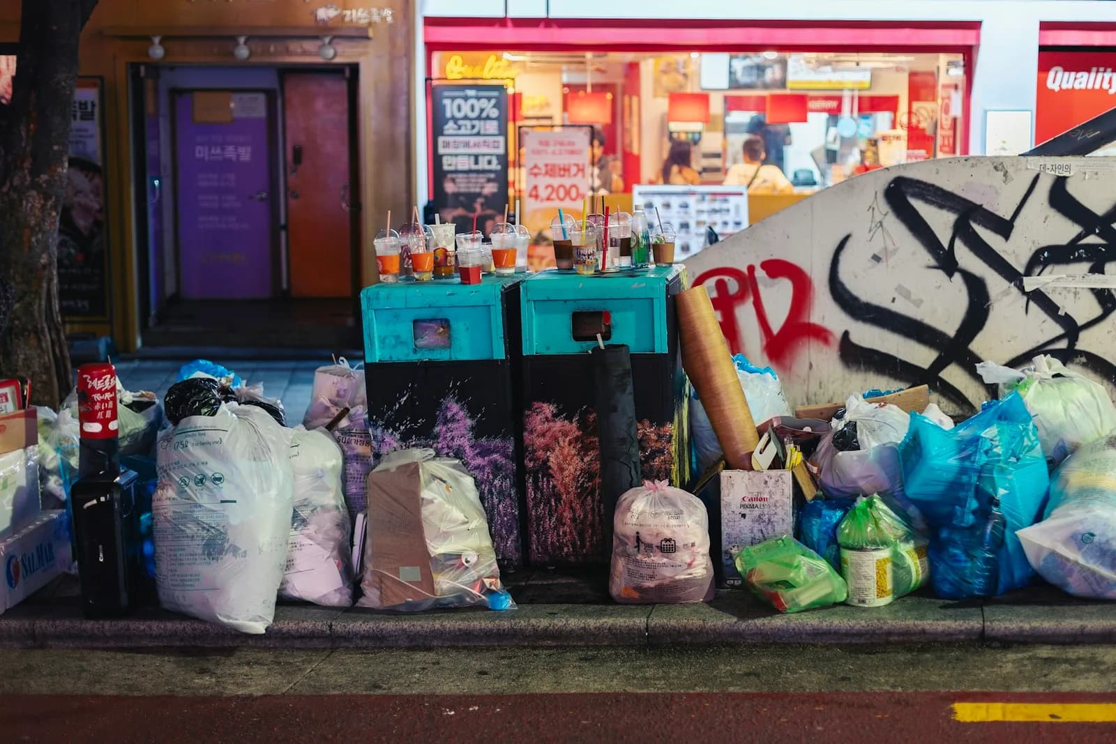 Sorted household waste piled at the kerb on a Hongdae street at night: white district bags, blue and clear recycling bags, flattened cardboard