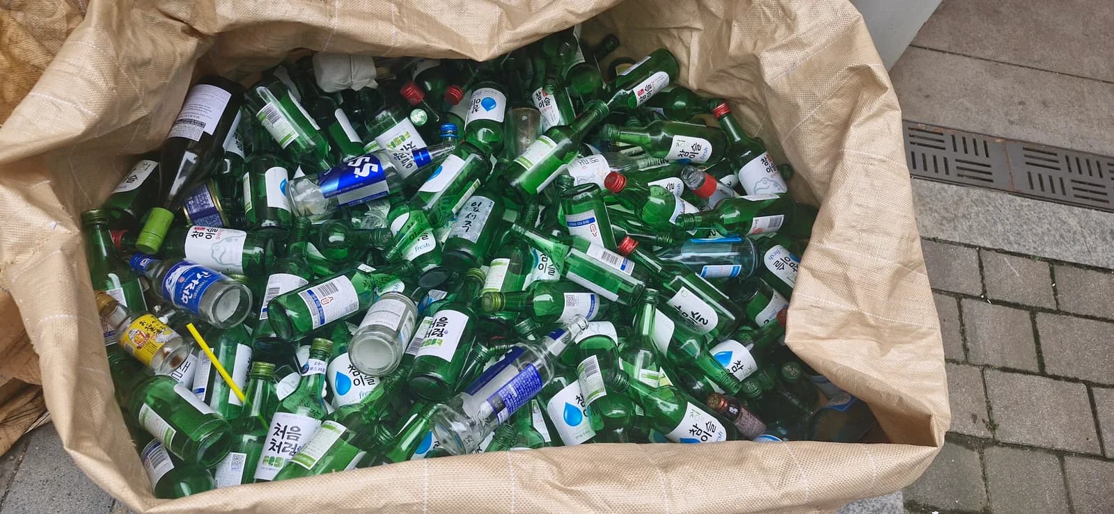 Empty green soju bottles collected in a large bag at a glass recycling point beside an apartment complex in Korea