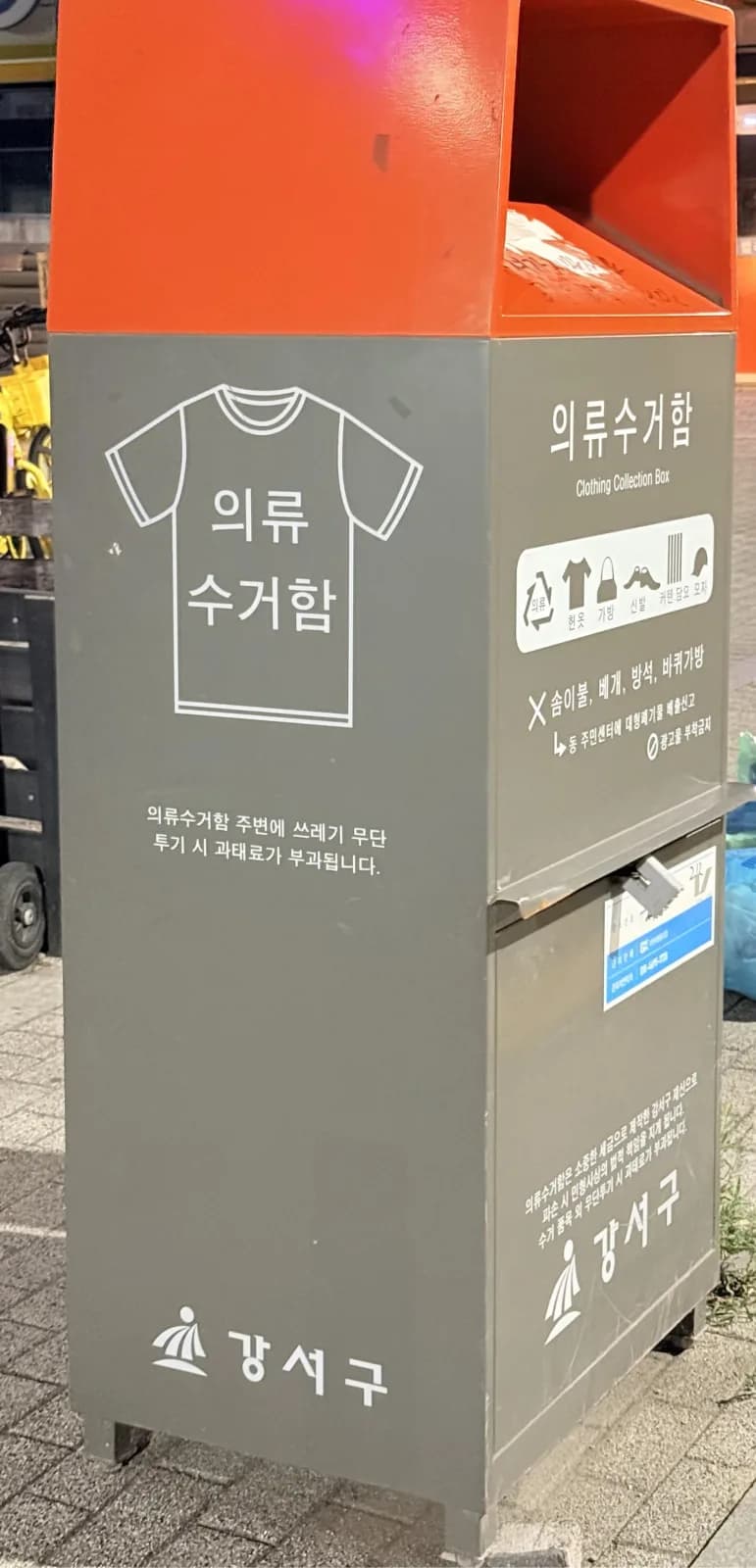 A Gangseo-gu clothing collection box at night: orange lid, grey body, a T-shirt pictogram, and icons for clothes, bags, shoes, curtains, blankets and hats