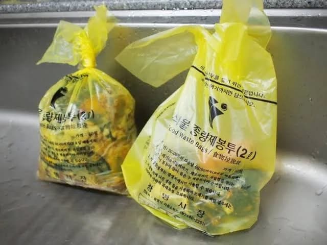 Two tied yellow 2-litre food-waste bags in a kitchen sink, the print reading 음식물 종량제봉투 2L