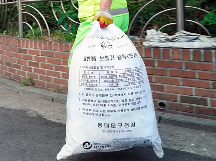 A collection worker carrying a white 75-litre Dongdaemun-gu general waste bag; the print lists collection days, the 6 pm to 9 pm window, and the fine for using any other bag