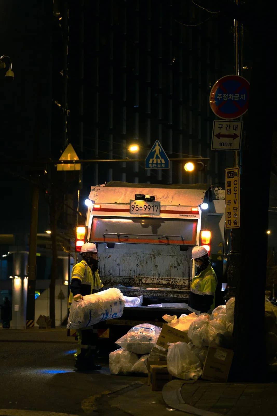 A refuse truck at night in Seoul with two workers loading white bags
